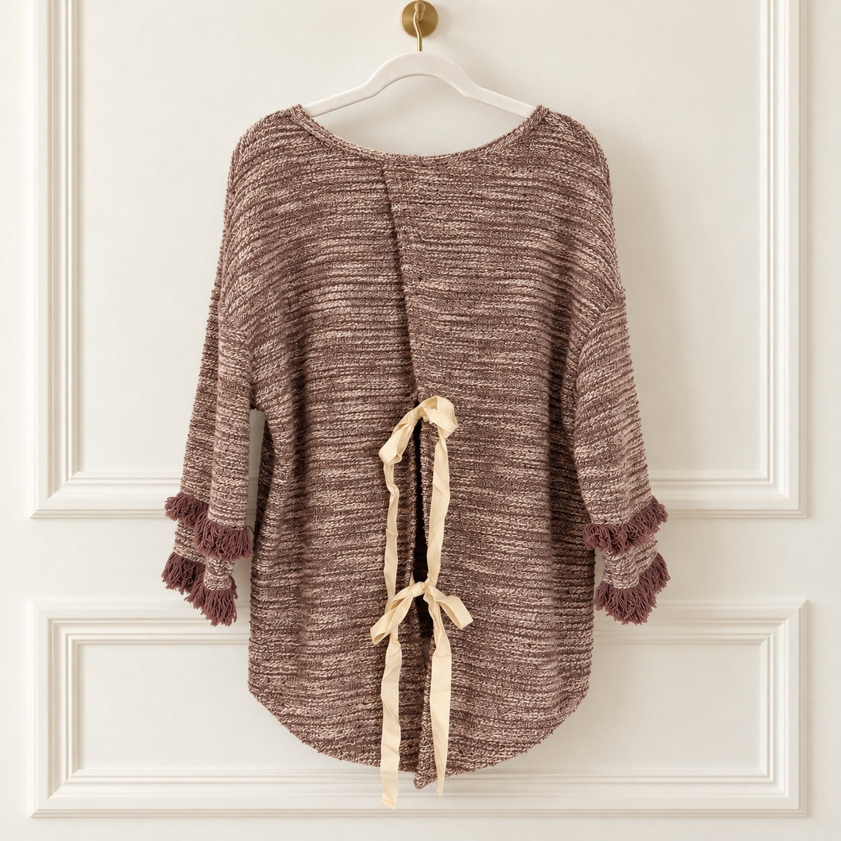 A brown and beige marled knit poncho-style top with fringe trim on the sleeves, hanging on a white hanger against a white paneled wall. The back has a tie detail with cream ribbon.