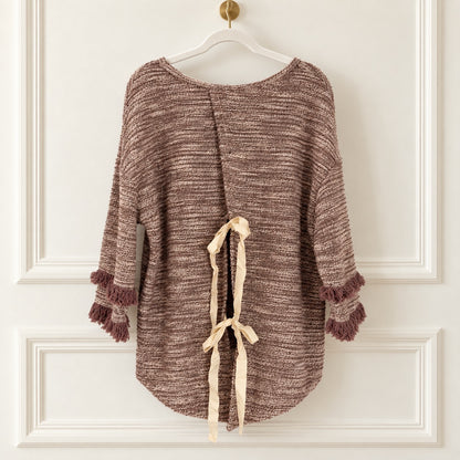 A brown and beige marled knit poncho-style top with fringe trim on the sleeves, hanging on a white hanger against a white paneled wall. The back has a tie detail with cream ribbon.