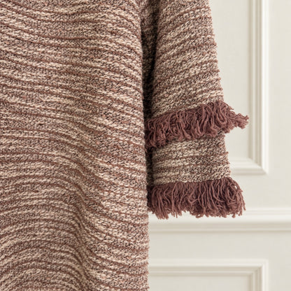 Close-up of a textured knit blanket or throw in heathered brown and beige tones, with fringed trim, shown against an off-white paneled wall.