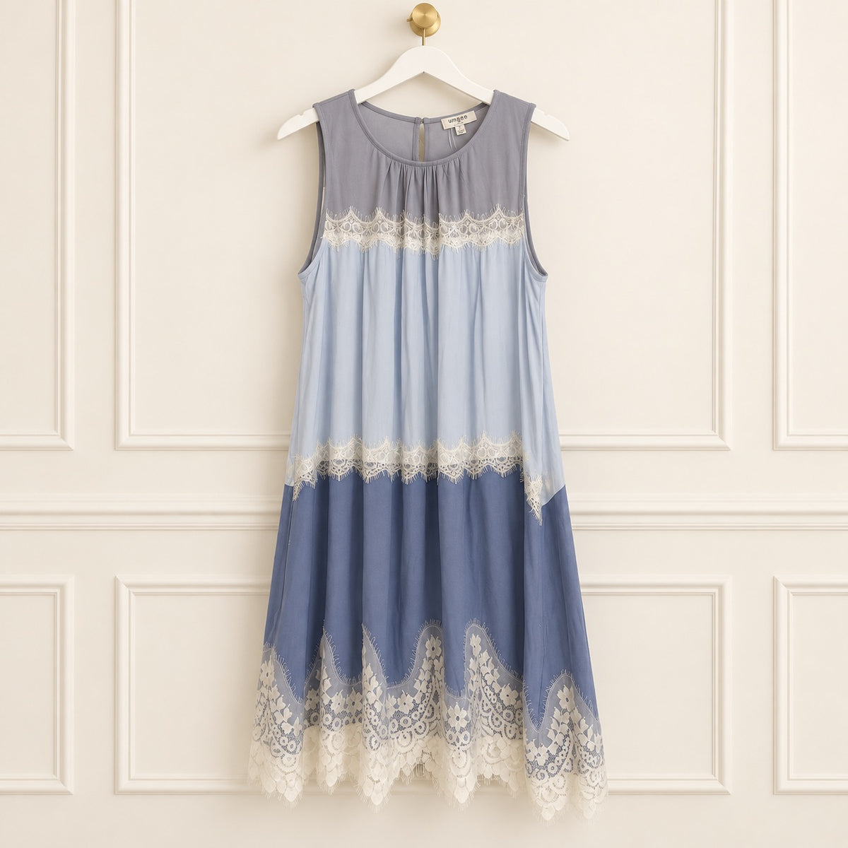 A sleeveless midi dress hanging on a white hanger against a cream paneled wall. The dress features a color-blocked design in slate gray, light blue, and dusty blue, with white lace trim at the chest, mid-skirt, and hem.