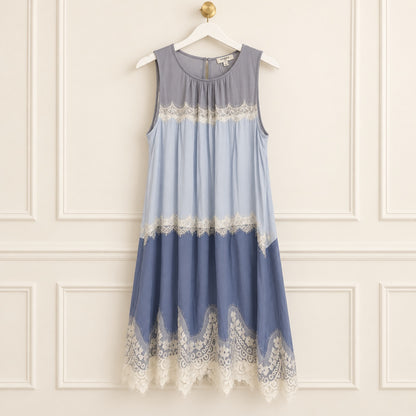 A sleeveless midi dress hanging on a white hanger against a cream paneled wall. The dress features a color-blocked design in slate gray, light blue, and dusty blue, with white lace trim at the chest, mid-skirt, and hem.