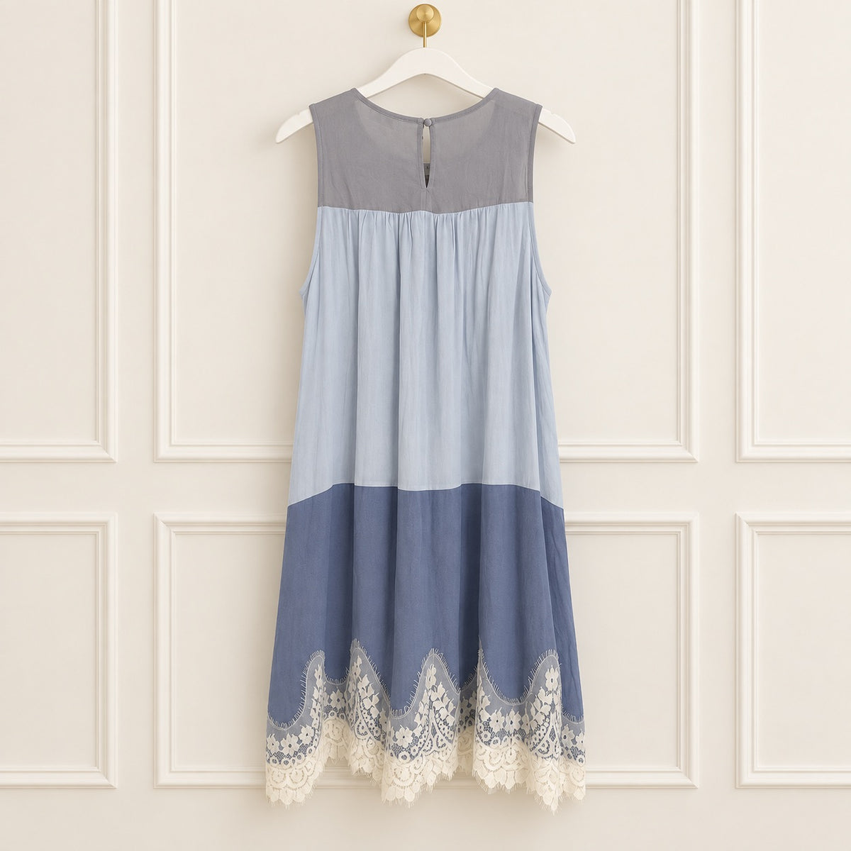 A sleeveless color-block midi dress in shades of gray, light blue, and medium blue, finished with a cream lace hem, hanging on a white hanger against a cream paneled wall.