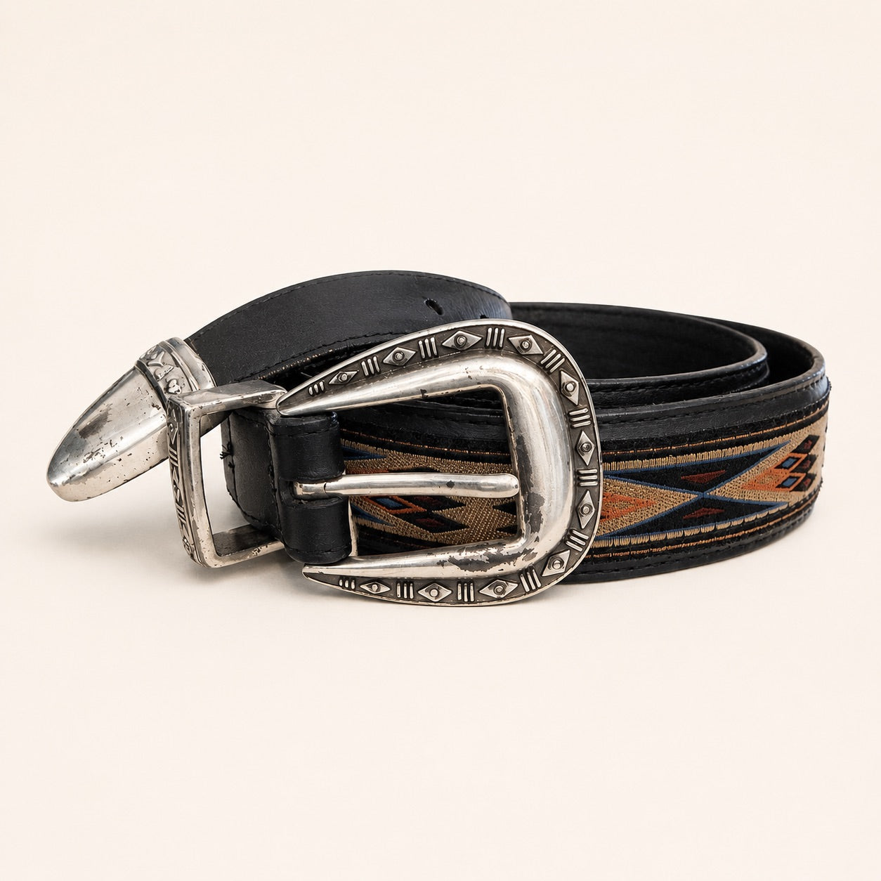 A western-style leather belt with a black strap, a silver-toned engraved buckle, and a woven geometric pattern in brown, orange, and blue, displayed on a light beige background.